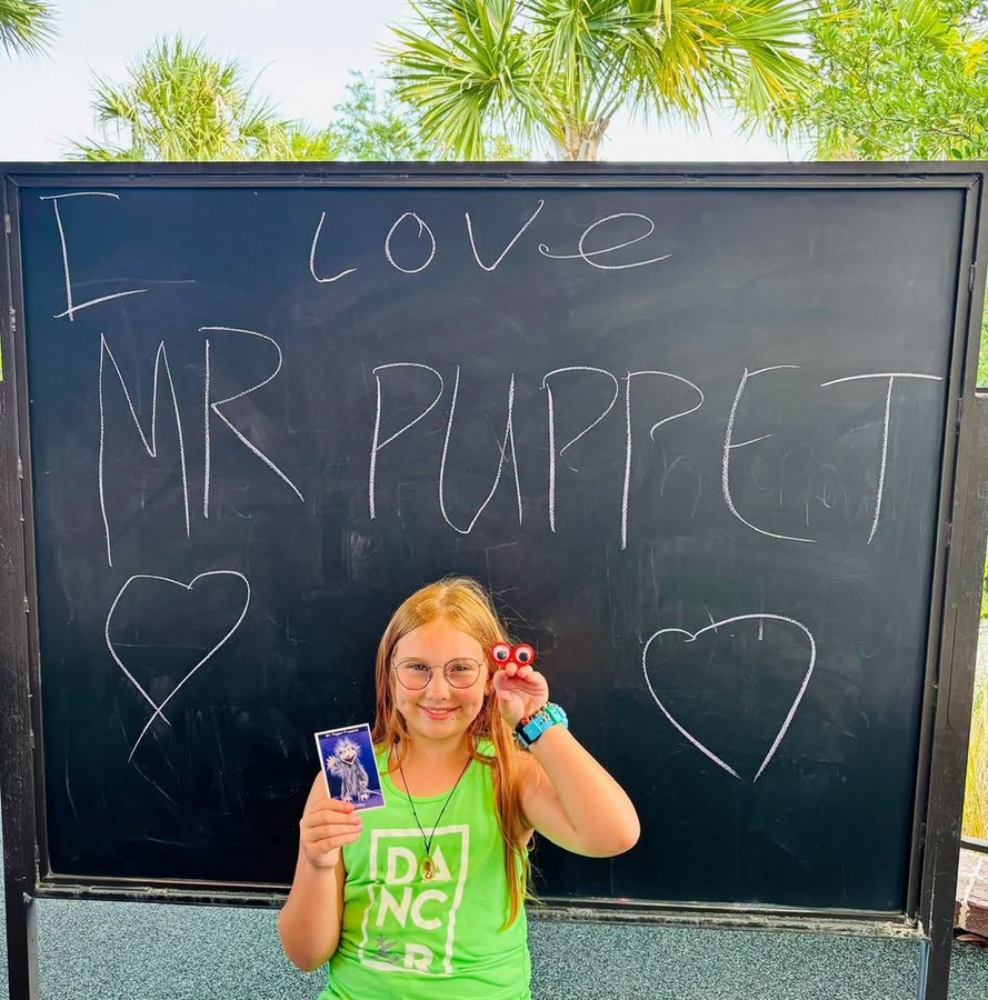 Smiling child holding a red peeper puppet in front of a chalkboard that reads I LOVE MR PUPPET